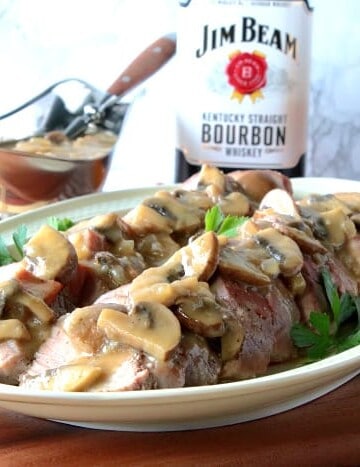 A sliced pork tenderloin on a platter with mushroom gravy and a glass of bourbon in the background.