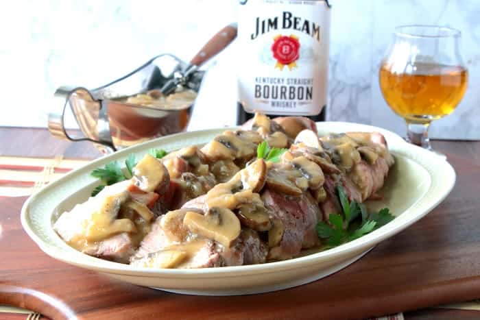 A sliced pork tenderloin on a platter with mushroom gravy and a glass of bourbon in the background.