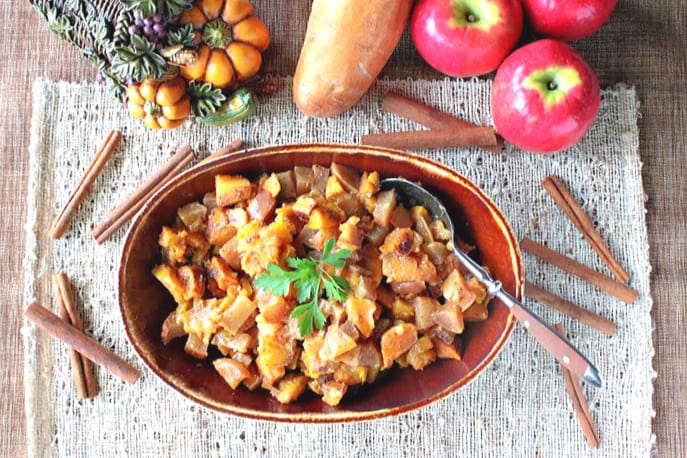 Overhead photo of a bowl of cinnamon sweet potatoes with cinnamon sticks, apples and a sweet potato in the background.