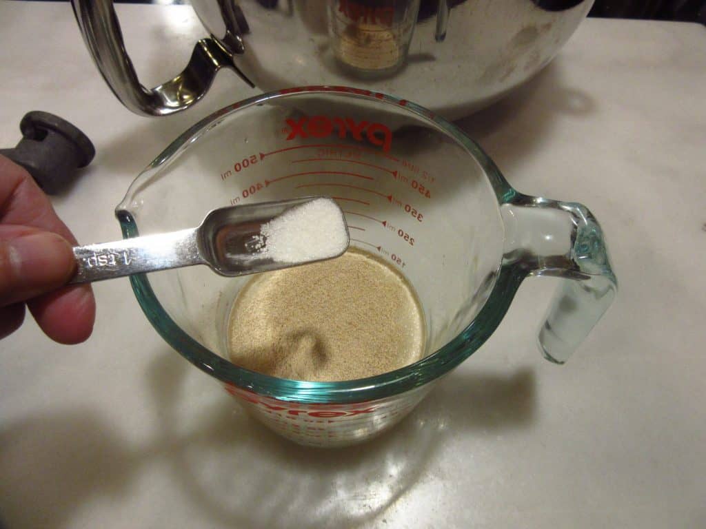 A touch of sugar being added to a bowl of yeast and water.