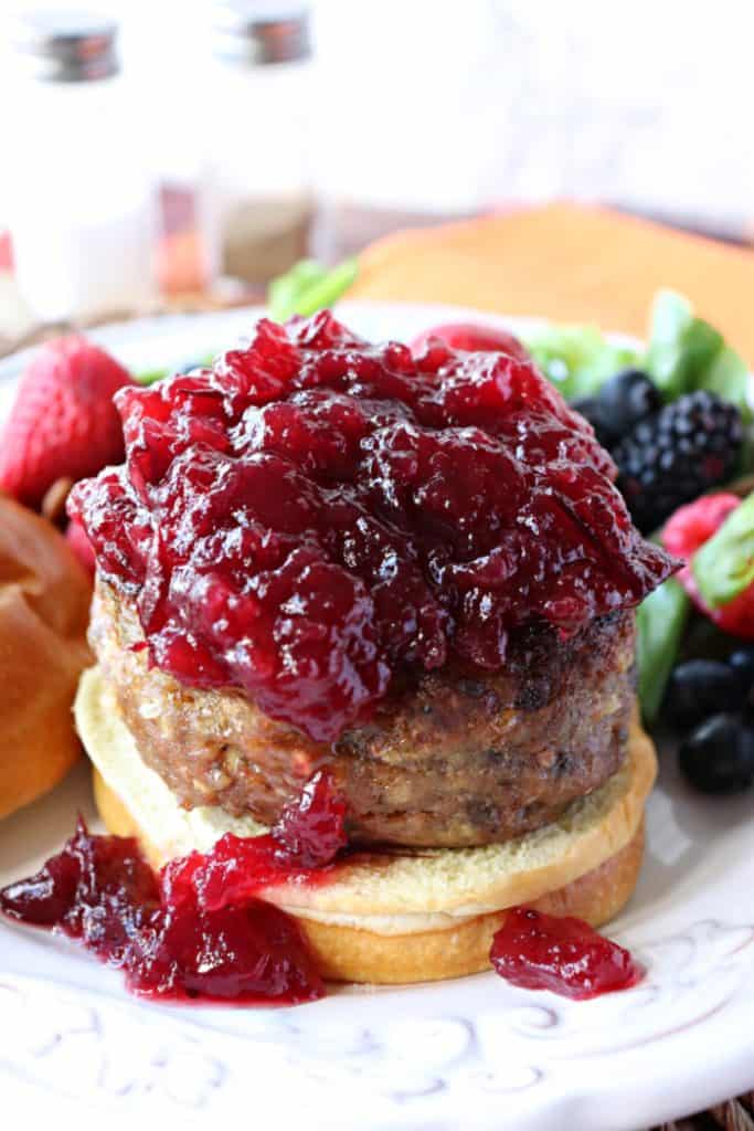 A vertical photo of a turkey burger topped with lots of cranberry sauce on a white plate.