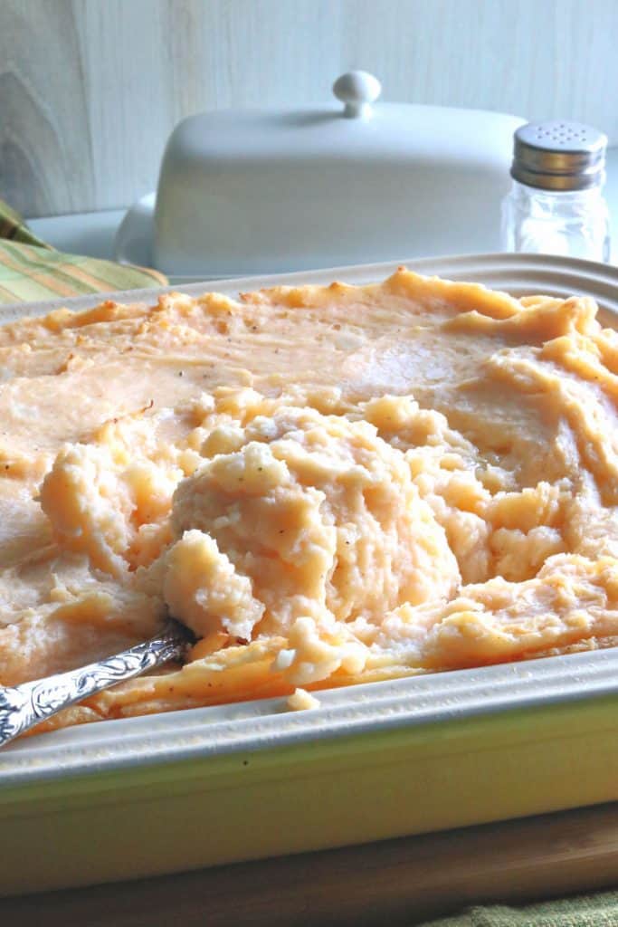 A vertical closeup photo of a casserole dish filled with cheesy horseradish mashed potato casserole with a serving spoon.