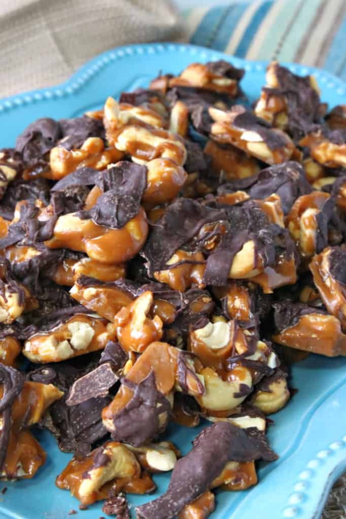 Closeup vertical photo of a pile of toffee cashew crunch on a blue plate with a dark chocolate drizzle.