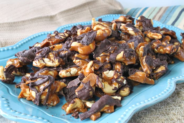 Horizontal photo of a platter of butter toffee cashew crunch with a chocolate drizzle and a blue and tan striped napkin.