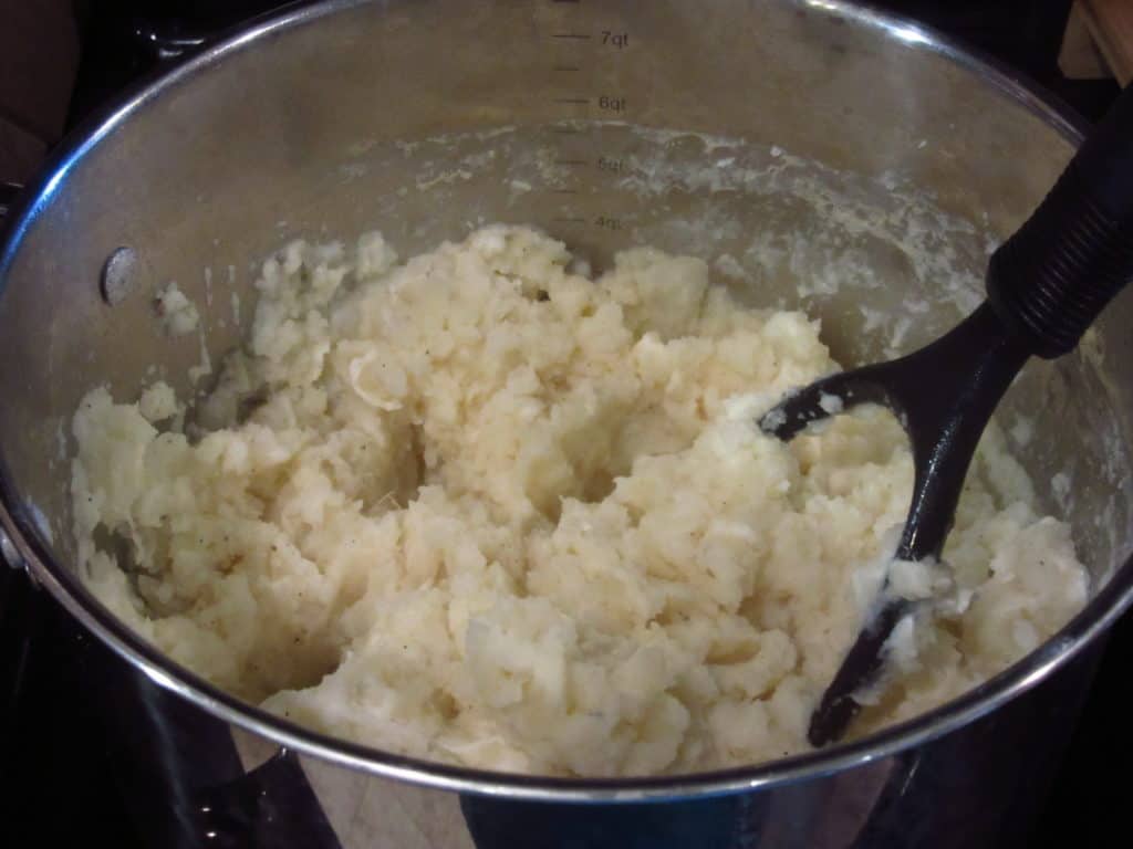 A potato masher in a pot of cooked potatoes.