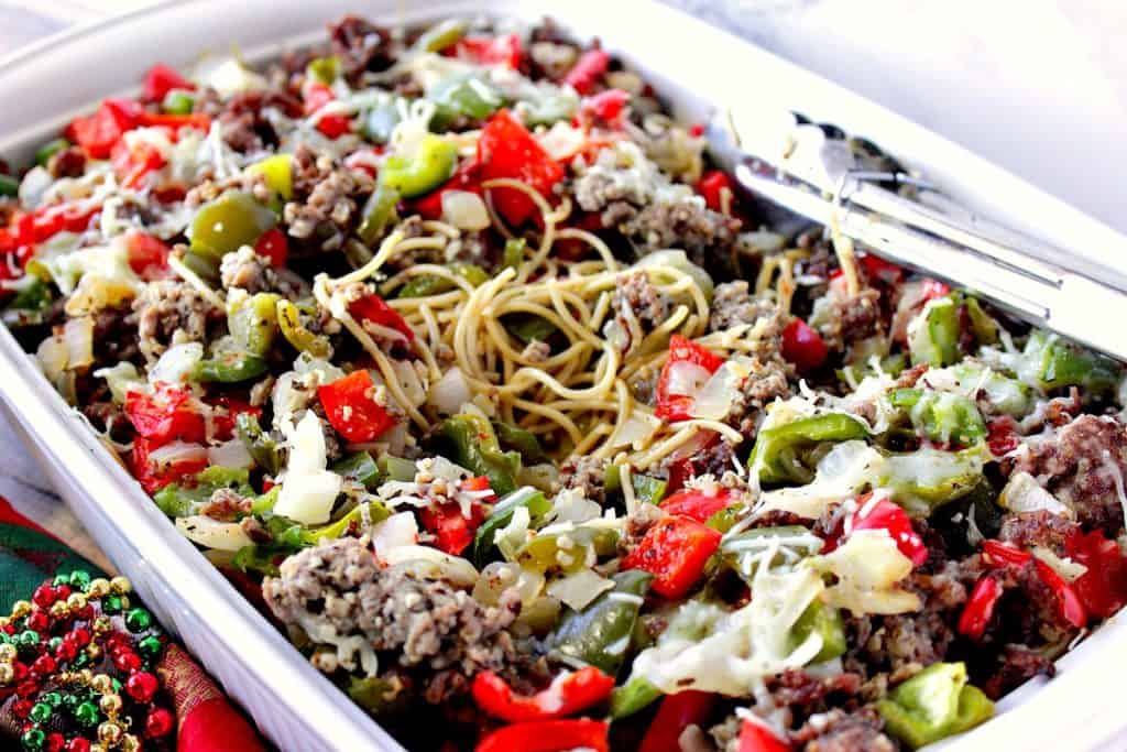 A horizontal closeup photo of spaghetti, sausage, and peppers in a casserole dish with tongs.