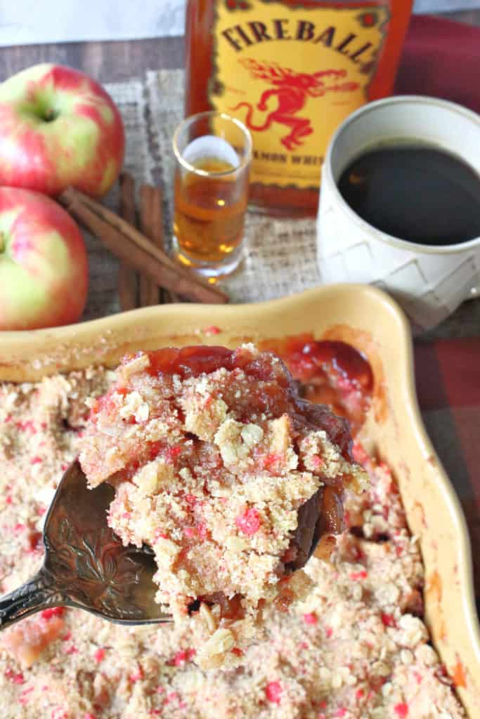 A vertical image of an apple crisp with cinnamon red hot topping, a shot of whiskey, cup of coffee, and apples.