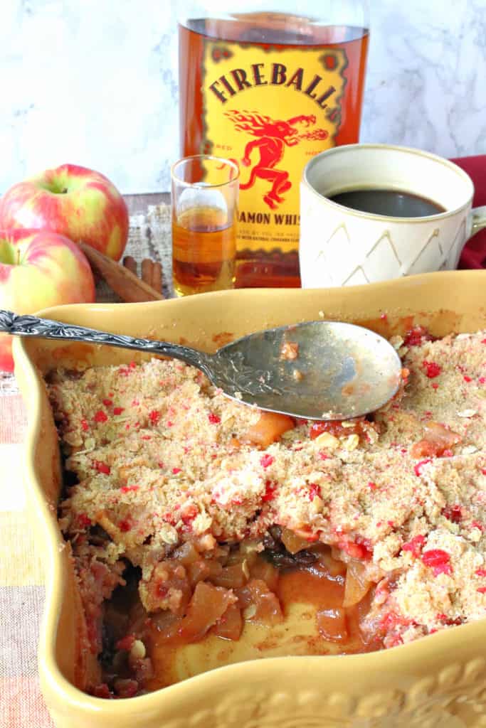 Vertical closeup photo of a fireball apple crisp with a serving taken out, a serving spoon, and a cup of coffee and cinnamon whiskey in the background.