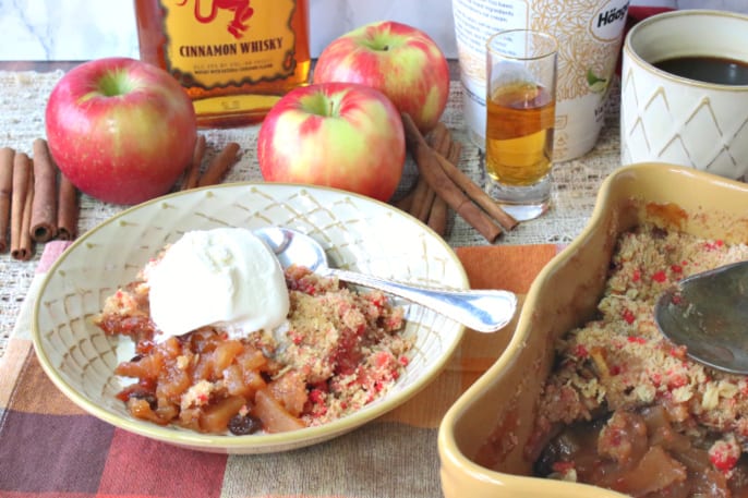 Horizontal photo of a bowl of fireball apple crisp with vanilla ice cream, apples, cinnamon sticks and a shot of cinnamon whiskey in the background.