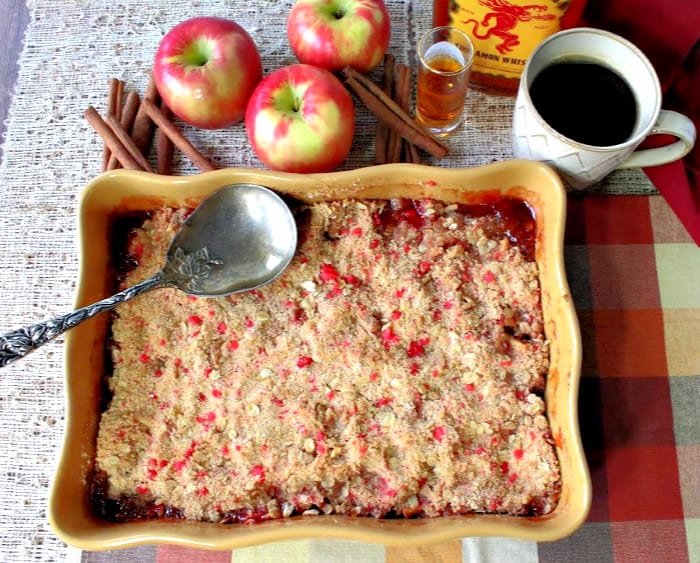 Overhead photo of a rectangle baking dish filled with fireball apple crisp with crushed red hot candy, a cup of coffee, serving spoon, apples, and cinnamon sticks.