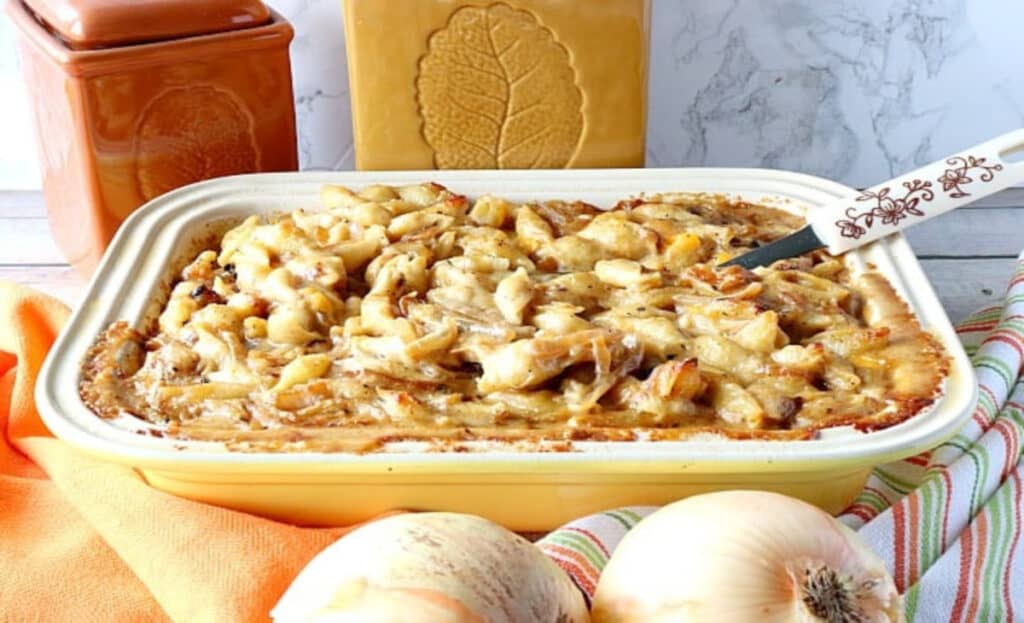A horizontal photo of French Onion Mac and Cheese in a casserole dish with a spoon and two onions in the forefront.