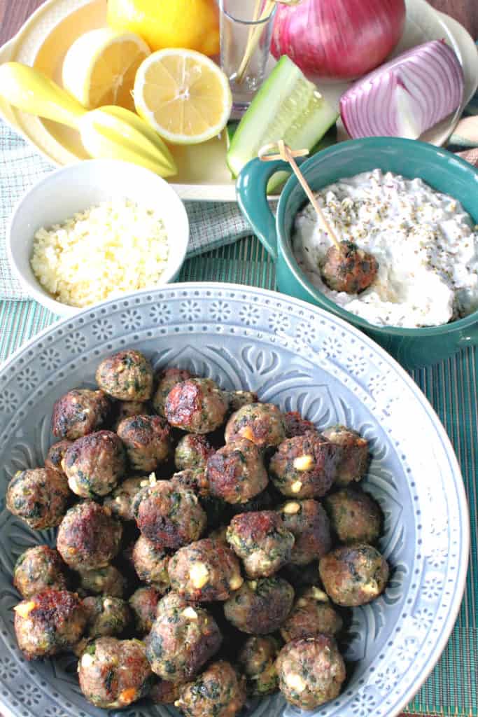 A vertical photo of a blue bowl of Greek appetizer meatballs with feta cheese, spinach, and tzatziki sauce.