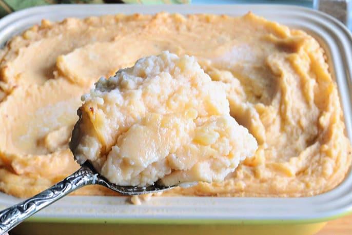 A closeup horizontal photo of a casserole dish filled with creamy, cheesy mashed potatoes on a serving spoon.