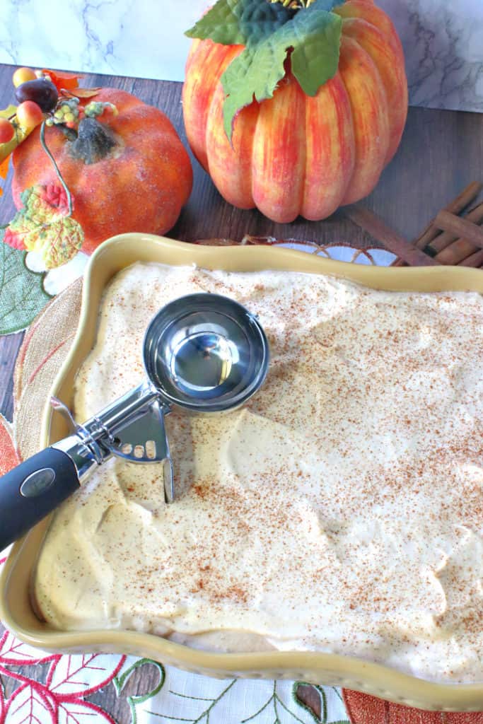 A vertical photo of a baking dish filled with pumpkin ice cream sprinkled with cinnamon and an ice cream scoop on top.