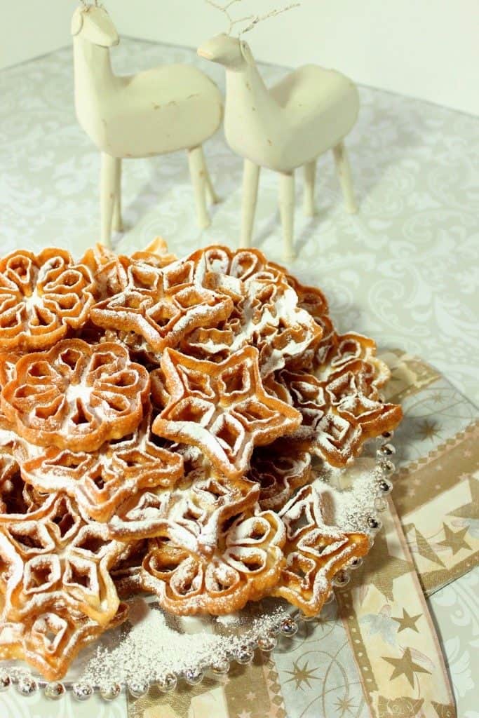 A vertical photo of a pile of rosette snowflake cookies on a glass plate with confectioners sugar and two white wood deer in the background.