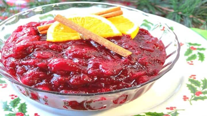 A glass bowl filled with Whole Berry Cranberry Sauce with cinnamon sticks and orange slices on top.