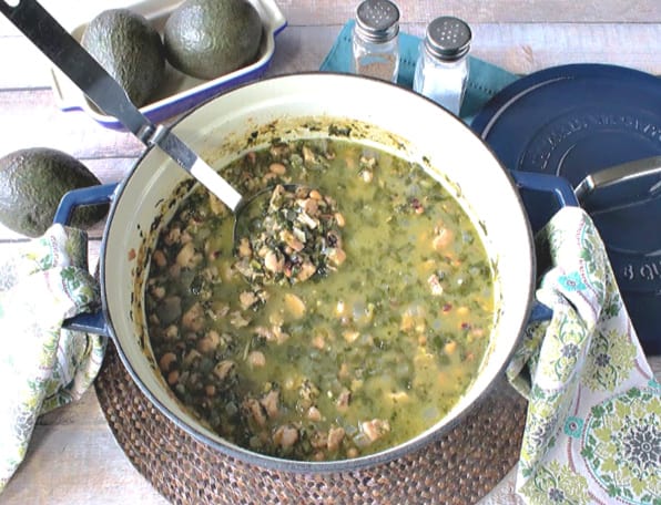 Overhead horizontal photo of a large pot of black-eyed pea chicken chili with a ladle, napkins, salt and pepper shaker, and avocados.