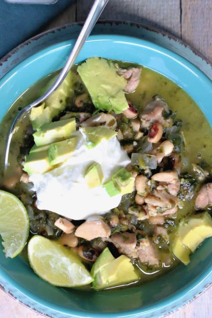 Closeup overhead vertical photo of a blue bowl filled with black-eyed pea chicken chili with avocado, lime, and sour cream.