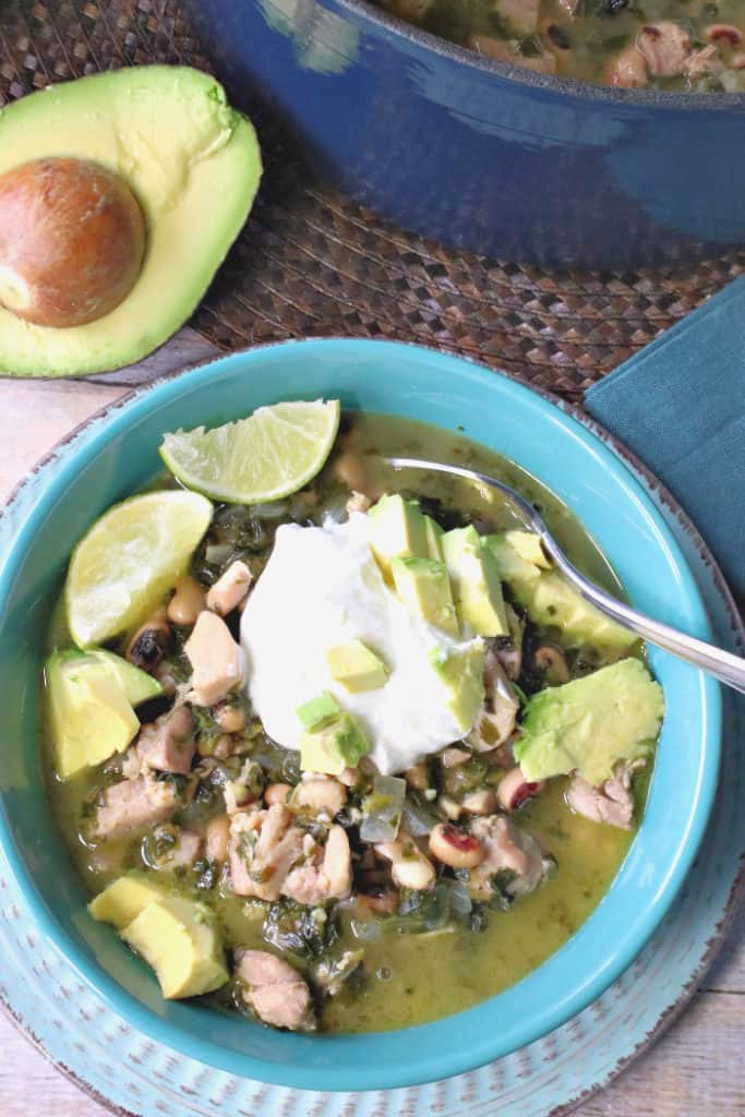 A vertical photo of a blue bowl filled with chicken chili with black-eyed peas, avocado, sour cream, and spinach.