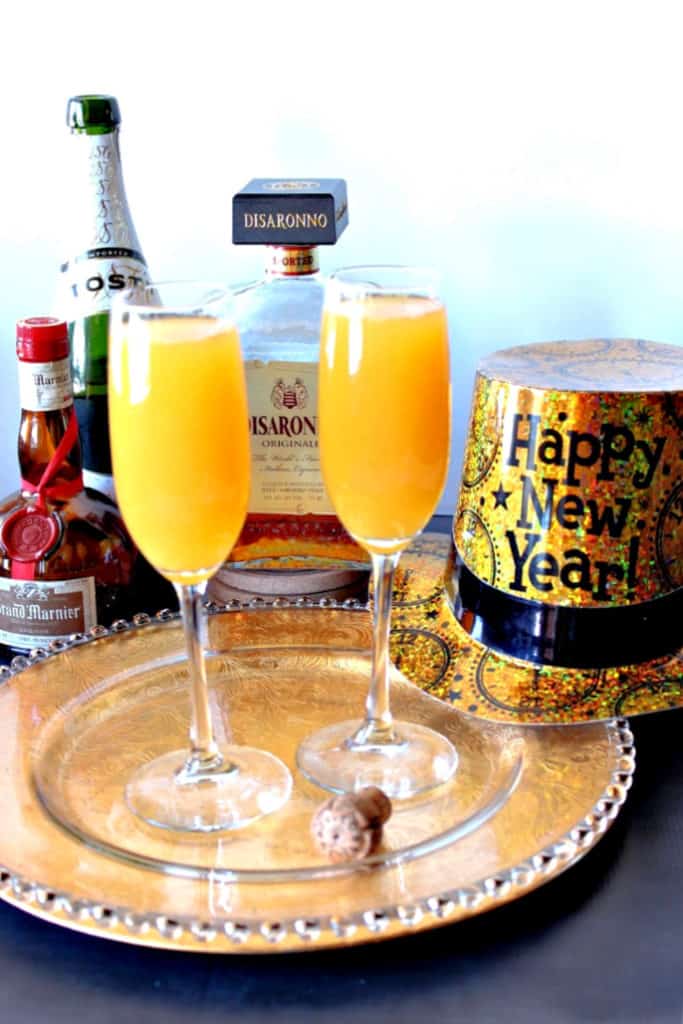 Two champagne glasses filled with the very best mimosas with amaretto, champagne and grand marnier in the background along with a new year's hat.