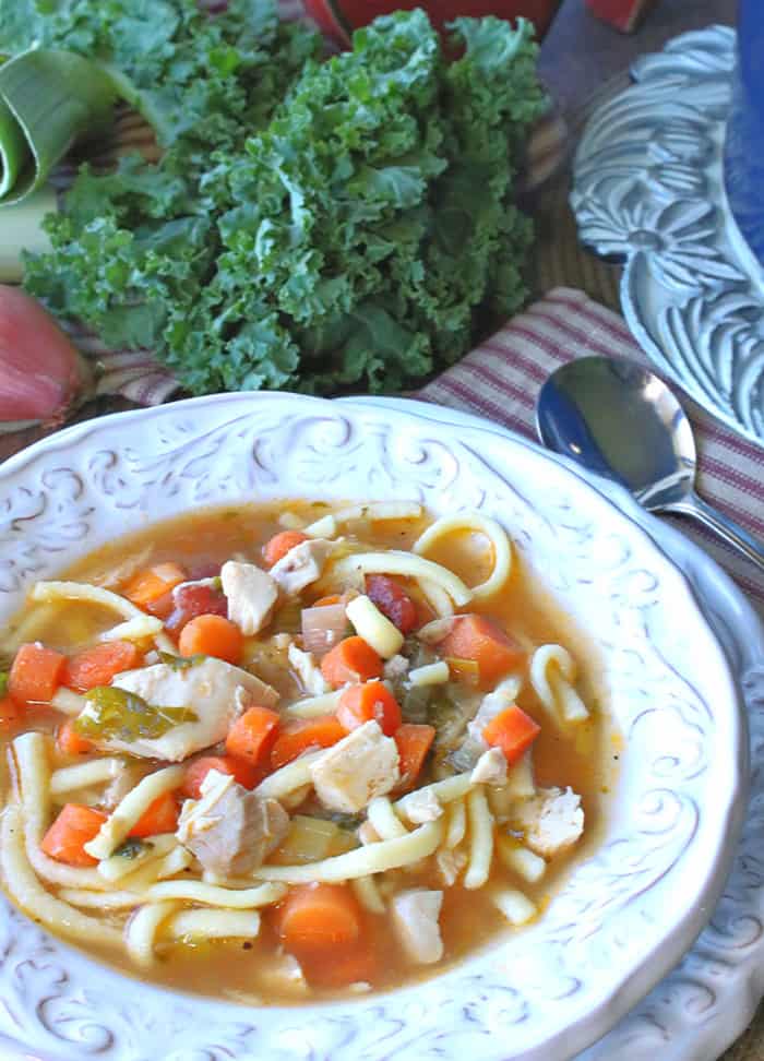 A closeup vertical photo of a inviting bowl of chicken and leek soup with carrots, and noodles with fresh kale in the background and a spoon on the side.