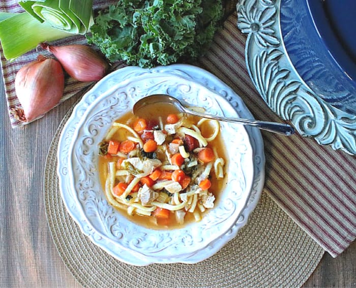 Overhead photo of a decorative white bowl filled with chicken and leek soup with a soup pot in the corner and leek, shallots, and kale in the background.