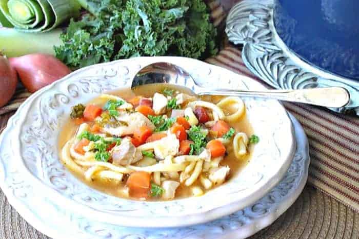 A horizontal photo of a bowl of chicken and leek soup with noodles, kale, carrots and a soup spoon.