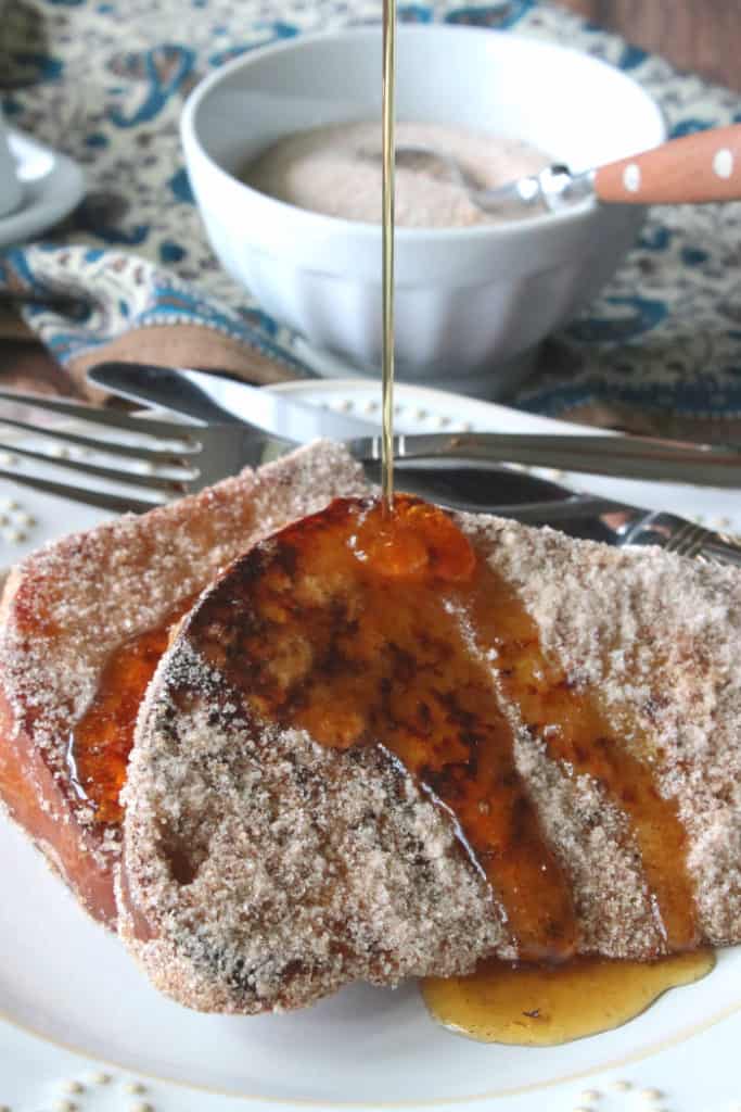 A closeup vertical photo of cinnamon sugar french toast on a plate with a stream of maple syrup being poured on top of it.