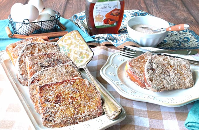 A horizontal photo of a rectangle plate filled with cinnamon sugar french toast with a serving spatula, a basket of eggs, and maple syrup in the background.