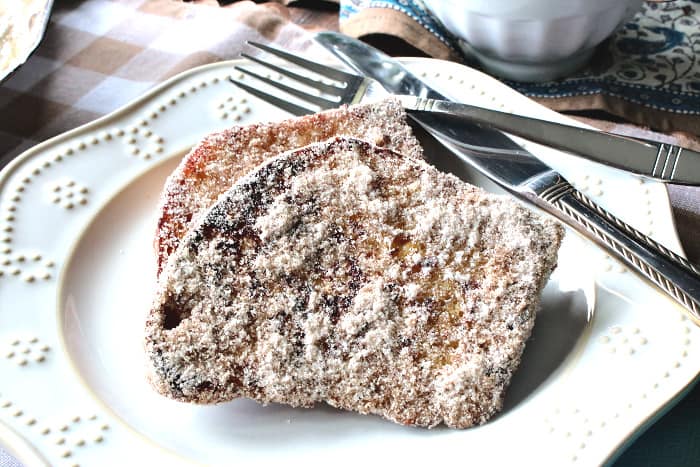 A decorative round plate with two slices of snickerdoodle French toast covered with cinnamon sugar and a fork and a knife.