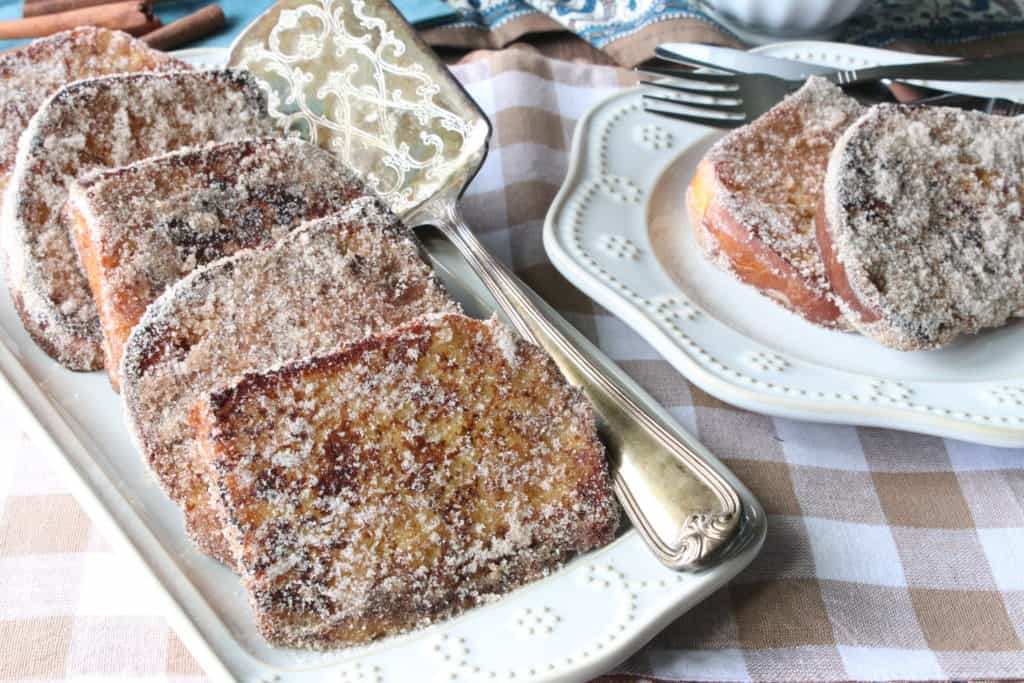 A horizontal photo of cinnamon sugar french toast on a rectangle plate with a serving spatula and a round plate with a fork. Both on a tan and white checked tablecloth.