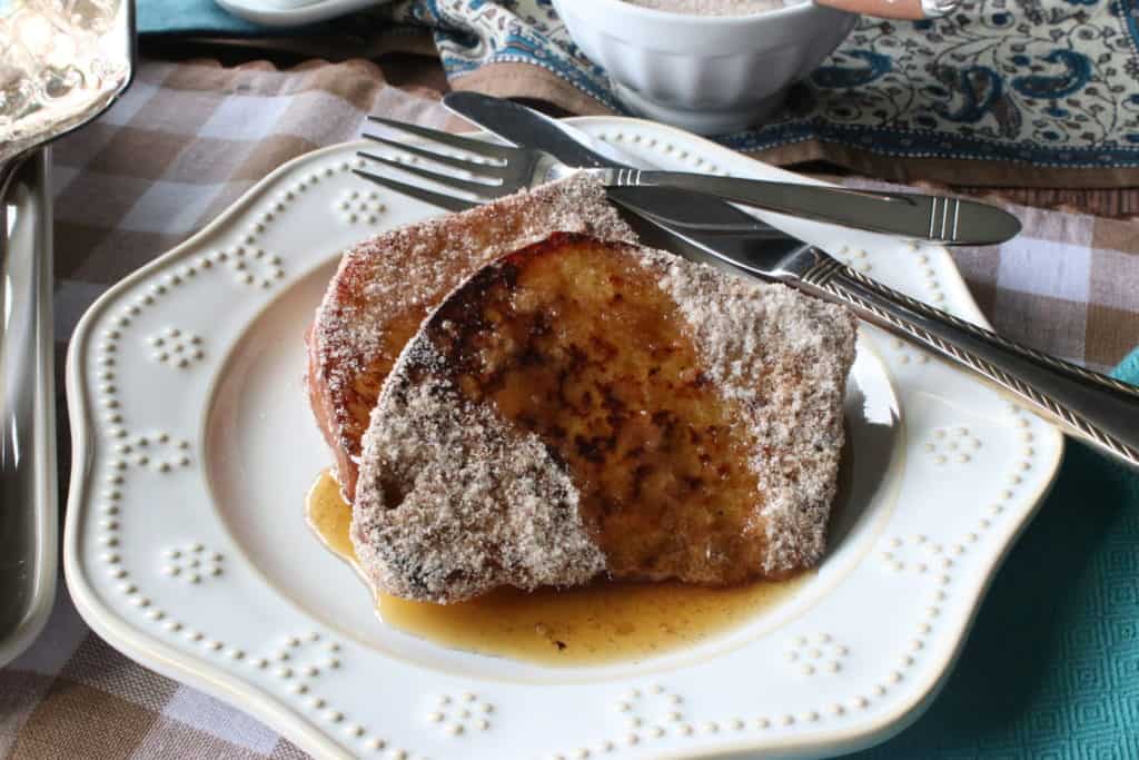 Two slices of cinnamon and sugar french toast on a pretty white plate with a fork, knife, and maple syrup.