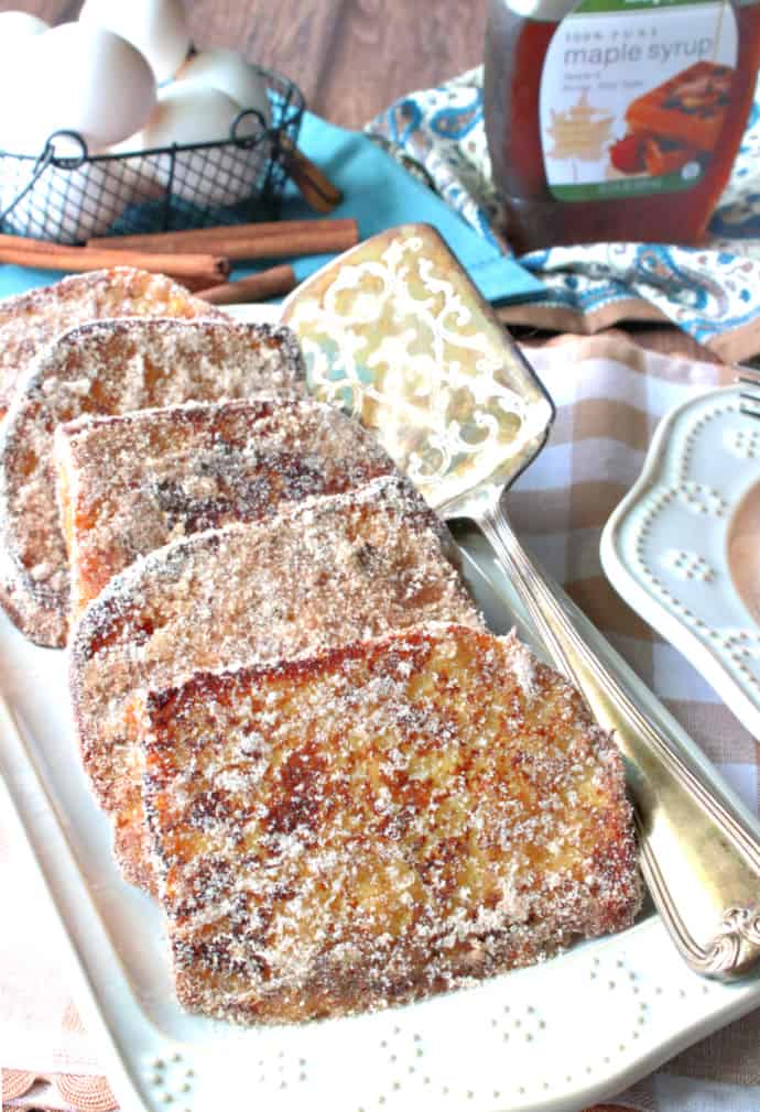 Vertical closeup image of a rectangle platter filled with cinnamon and sugar french toast with a basket of eggs in the background and a bottle of maple syrup.