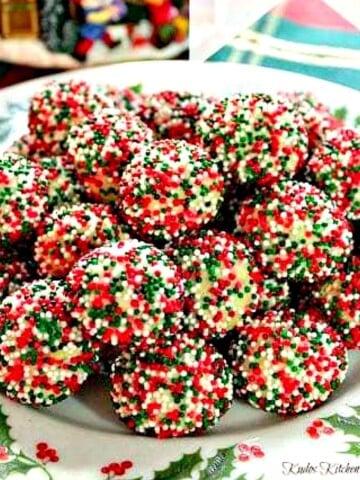 A holly berry plate stacked high with Christmas Cream Cheese Sprinkle Cookies.