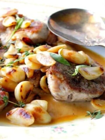 A platter filled with garlic lovers pork chops with whole garlic, sauce, fresh herbs and a serving spoon