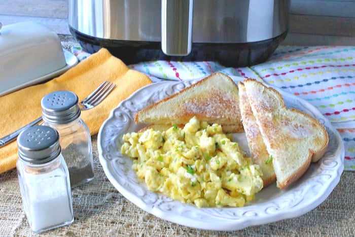 A white plate filled with air fryer scrambled eggs with chives. Toast and butter is on the side and salt and pepper shakers are in the foreground while and air fryer is in the background.