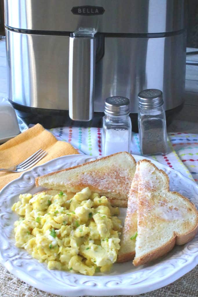 A closeup vertical photo of a plate of scrambled eggs with chives, toast with butter, and an air fryer in the background.