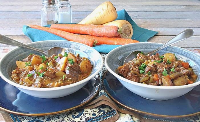 A horizontal photo of two side by side blue bowls filled with beef stew. Each are on a blue plate with spoons and carrots and parsnips are in the background.