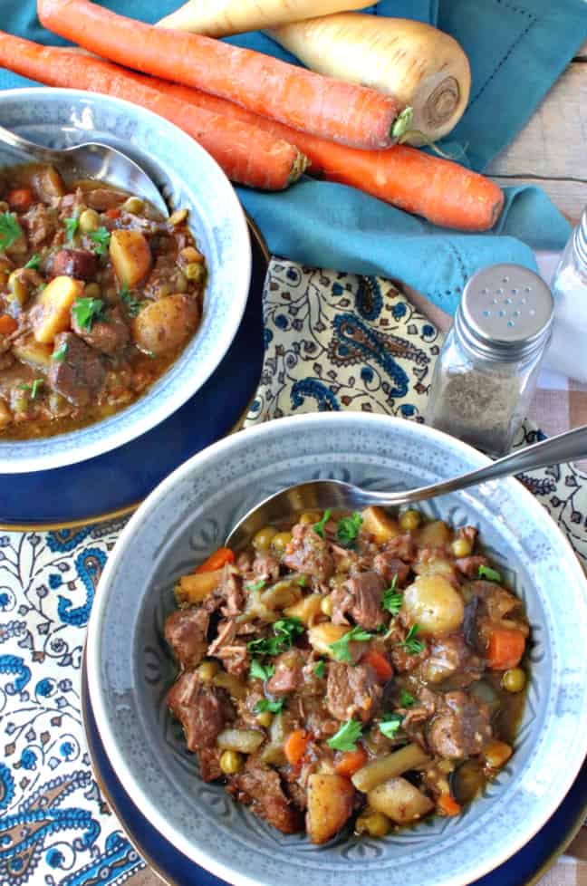 A vertical overhead photo of two blue bowls filled with delicious beef stew for a crowd with carrots, parsnips, peas, and beans.