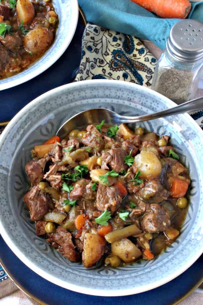 A closeup vertical image of a hot and hearty bowl of beef stew with a spoon, parsley, peas, potatoes, and pearl onions.