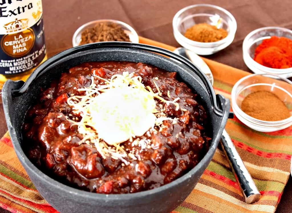 A horizontal photo of turkey chili in a small cast iron pot with small bowls of spices and a spoon in the background.