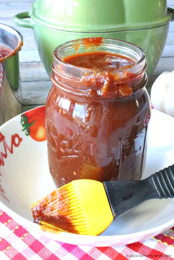 A vertical closeup image of salsa bbq sauce in a mason jar sitting in a bowl, with a sauce brush.