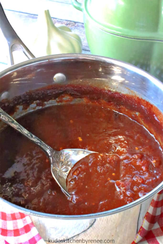 A vertical closeup image of a saucepan of homemade BBQ sauce made from salsa with a silver serving spoon.
