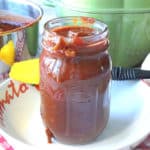 A jar of salsa BBQ sauce in a bowl with a brush and a saucepan in the background.