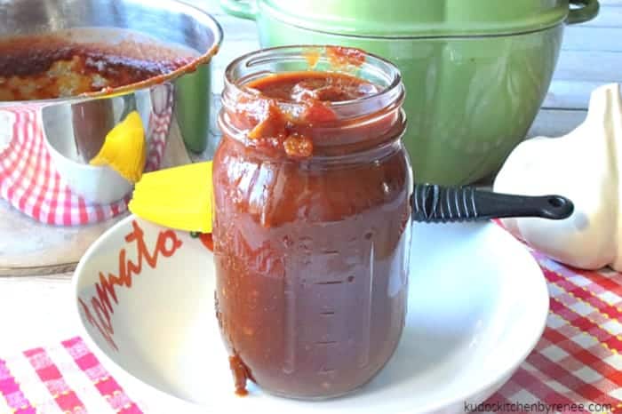 A horizontal photo of a mason jar filled with homemade bbq salsa on a red checked tablecloth with a saucepan and casserole dish in the background.