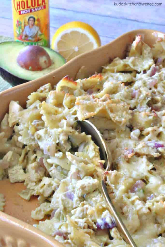 A vertical closeup image of a casserole dish filled with creamy chicken pasta with artichokes and avocados with a large serving spoon in the center.