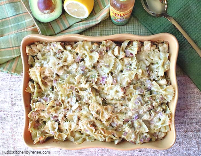 Overhead horizontal photo of a 9x13 casserole dish filled with creamy chicken and artichoke pasta with avocados with green napkins and a gold serving spoon.