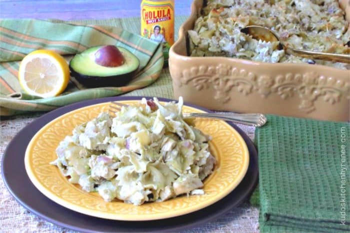 A horizontal photo of a plate of chicken pasta with artichokes and avocado with a casserole dish in the background along with a half a lemon and half an avocado.