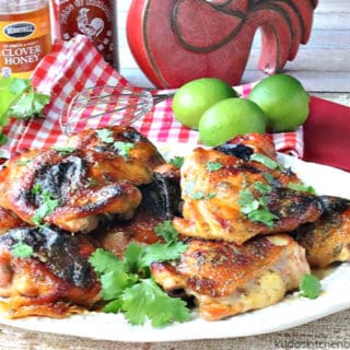 A horizontal photo of a platter of chicken thighs with honey lime sriracha sauce. Fresh limes, cilantro, honey and sriracha sauce is in the background.