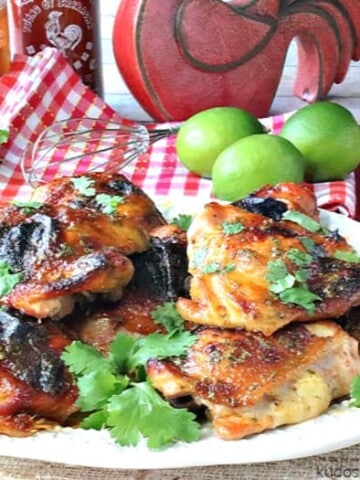 A horizontal photo of a platter of chicken thighs with honey lime sriracha sauce. Fresh limes, cilantro, honey and sriracha sauce is in the background.